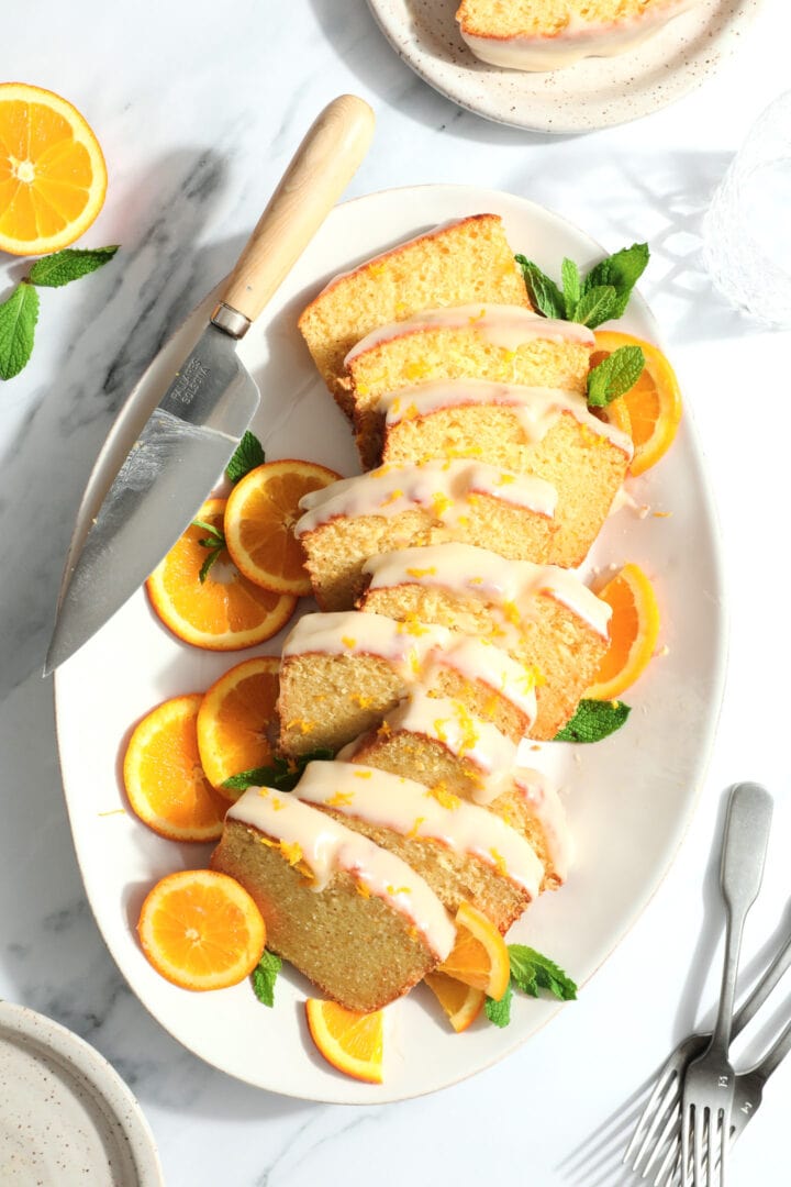 a sliced loaf cake on a white oval platter. The cake slices are glazed with a white frosting and sprinkled with orange zest.