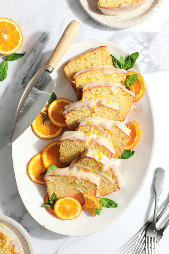 a sliced loaf cake on a white oval platter. The cake slices are glazed with a white frosting and sprinkled with orange zest.