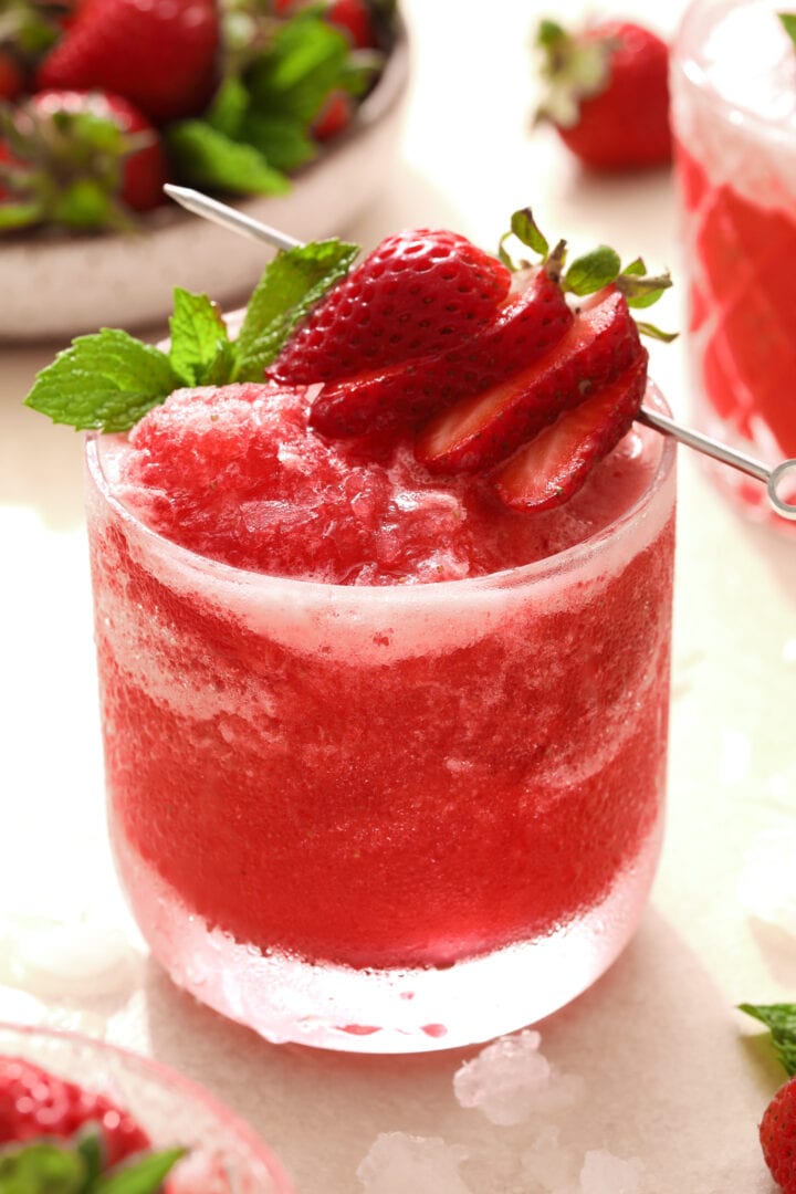 small glass of bright pink strawberry slush, garnished with fresh sliced strawberries and mint leaves.