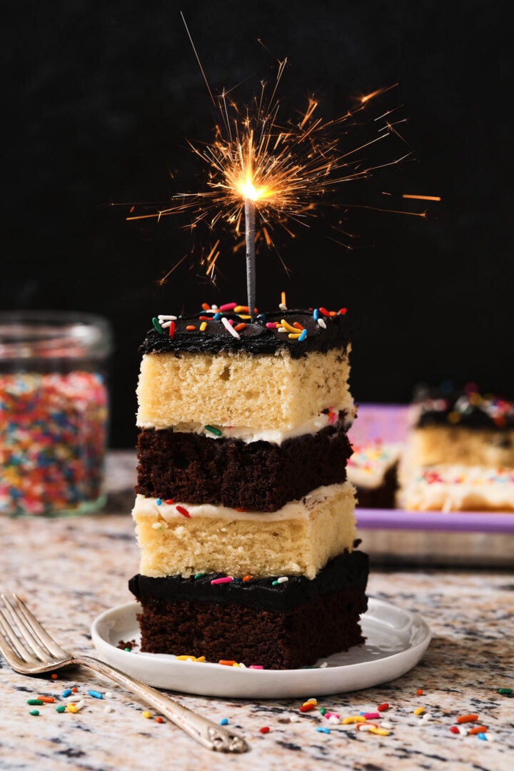 stack of chocolate and vanilla cake slices, alternating in a pattern between the chocolate and vanilla. There is a silver sparkler on top and a jar of sprinkles in the background to the left.