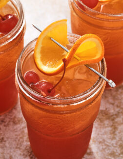 tall glass of fruity cocktail, garnished with a twisted orange slice and two maraschino cherries.