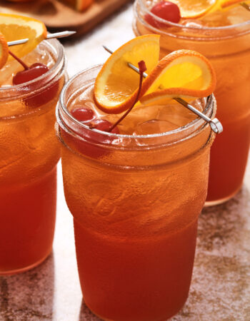 tall glass of fruity cocktail, garnished with a twisted orange slice and two maraschino cherries.