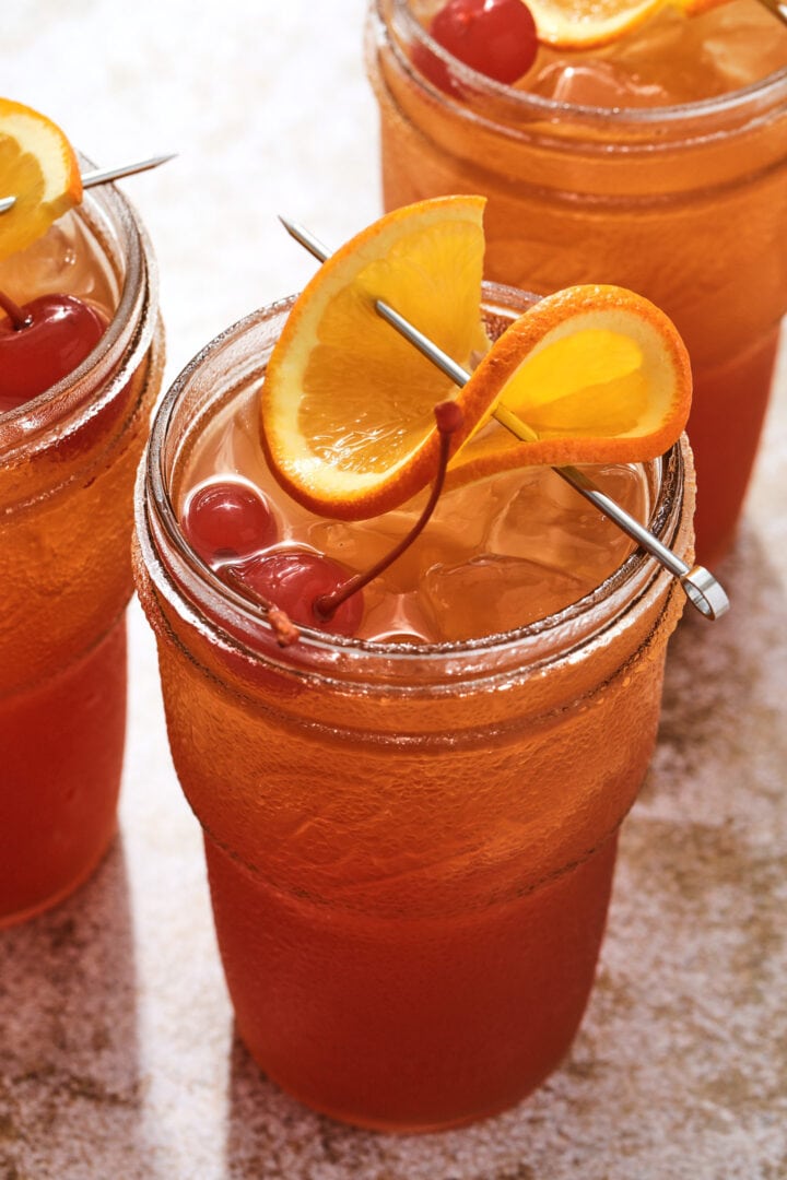 tall glass of fruity cocktail, garnished with a twisted orange slice and two maraschino cherries.