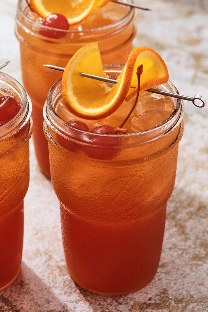 tall glass of fruity cocktail, garnished with a twisted orange slice and two maraschino cherries.