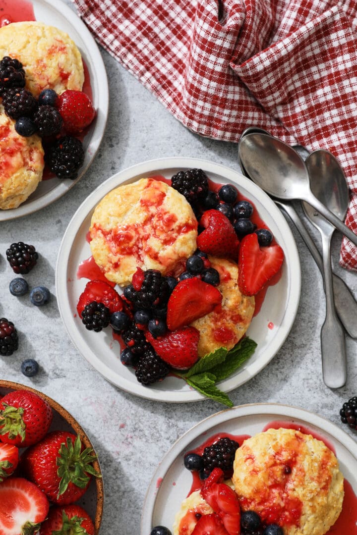 several small gray plates with biscuits, berries and whipped cream. There is a red checkered towel off to the left hand side.