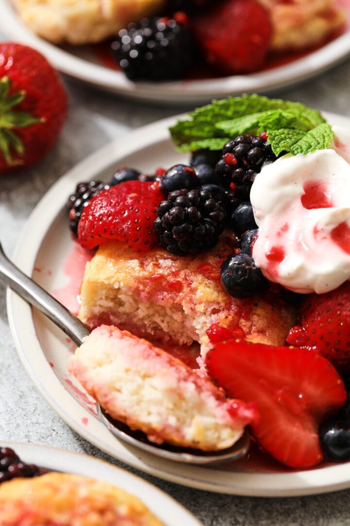 small gray plate of biscuits, berries and whipped cream. A sprig of mint leaves are near the top right hand corner and there is a strawberry off to the left.