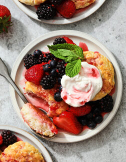 small gray plate of biscuits, berries and whipped cream. A sprig of mint leaves are near the top right hand corner and there is a strawberry off to the left.