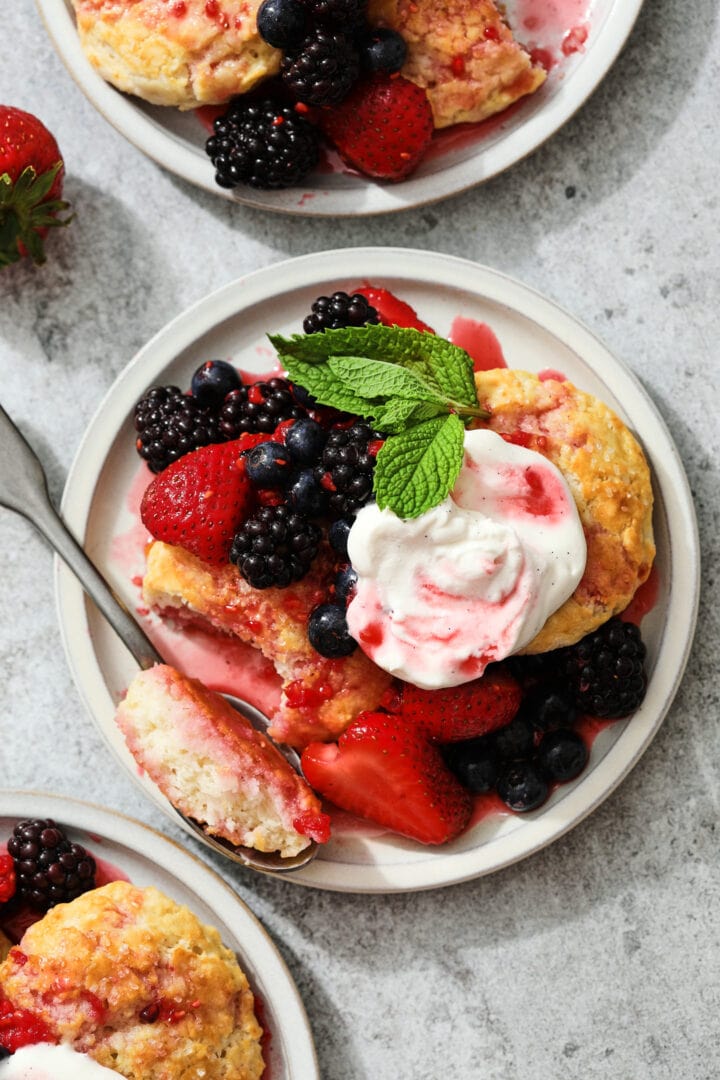 small gray plate of biscuits, berries and whipped cream. A sprig of mint leaves are near the top right hand corner and there is a strawberry off to the left.