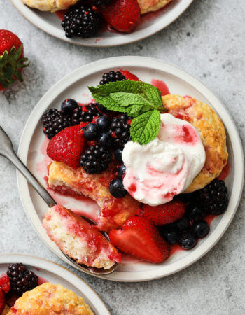 small gray plate of biscuits, berries and whipped cream. A sprig of mint leaves are near the top right hand corner and there is a strawberry off to the left.