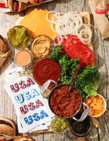 board of abundant condiments for hot dogs and hamburgers, including ketchup, mustard, pickle relish, sliced pickles, burger sauce, shredded cheese, chili, onion jam, lettuce, onion, tomatoes and sliced cheese.