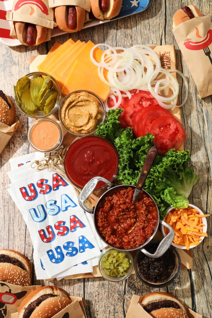 board of abundant condiments for hot dogs and hamburgers, including ketchup, mustard, pickle relish, sliced pickles, burger sauce, shredded cheese, chili, onion jam, lettuce, onion, tomatoes and sliced cheese.