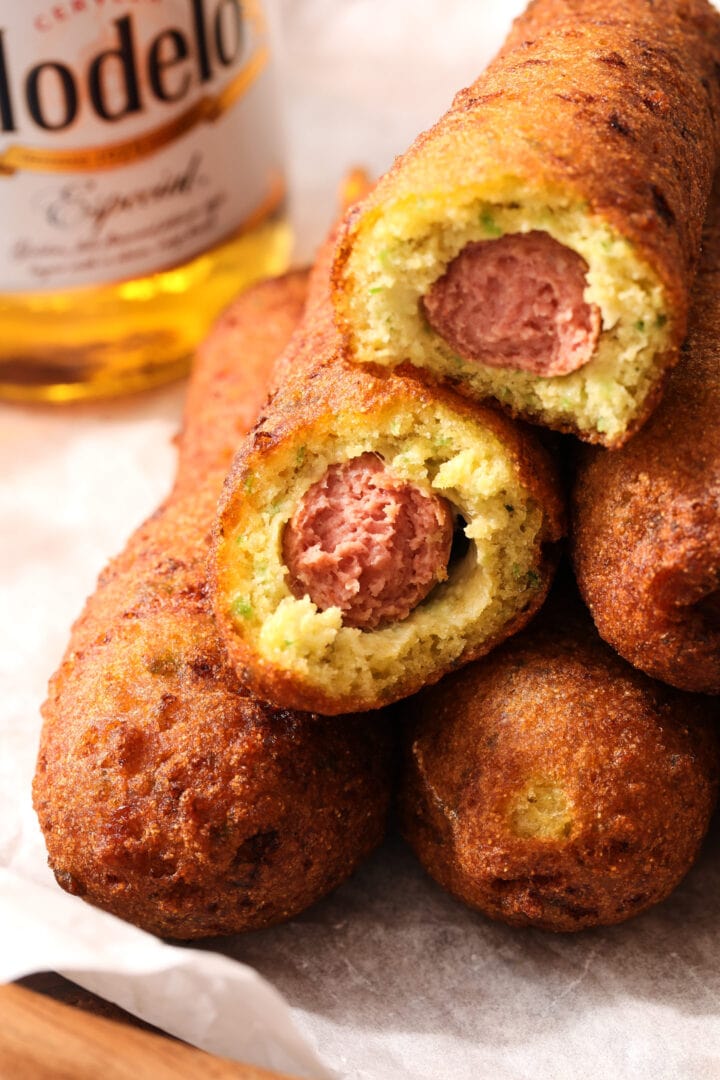 pile of corn dogs with a small bottle of beer in the background. Two of the corn dogs have bites taken out.