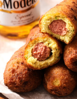 pile of corn dogs with a small bottle of beer in the background. Two of the corn dogs have bites taken out.