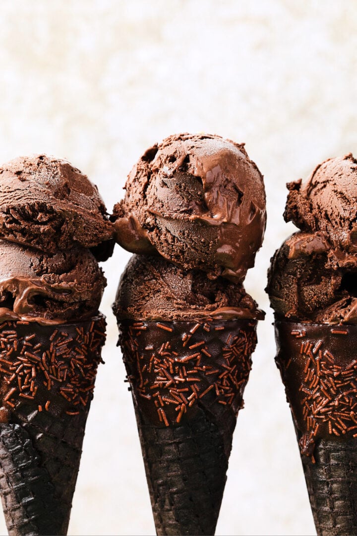three chocolate ice cream cones dipped in chocolate and sprinkles, with two scoops of chocolate ice cream on each cone.