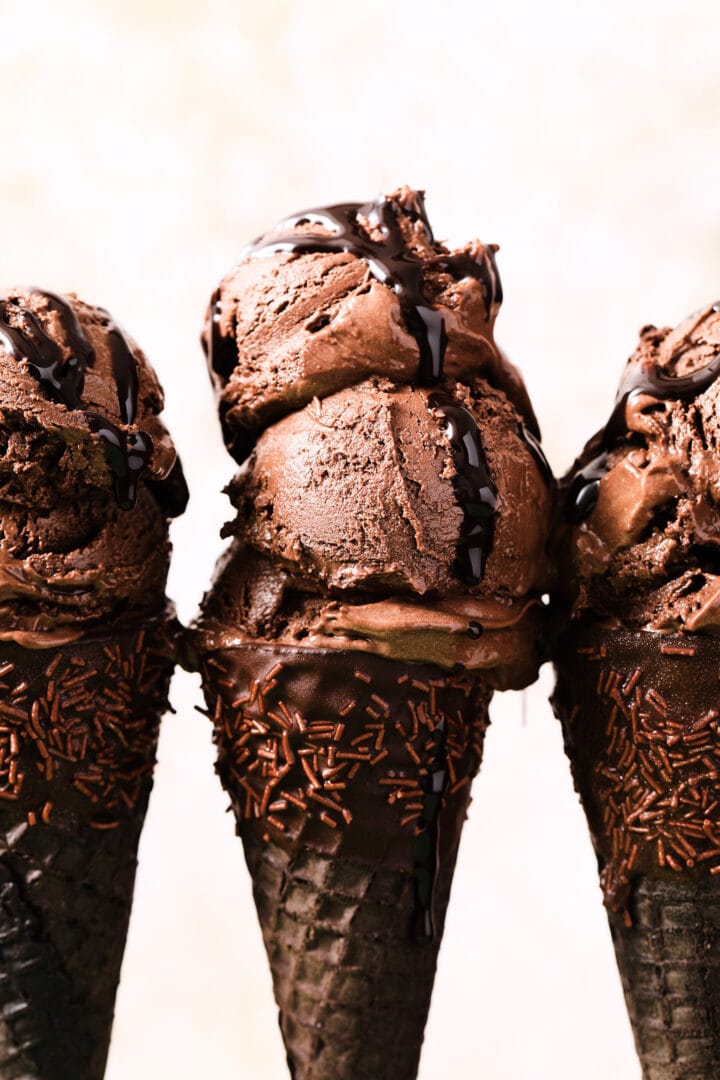 three chocolate ice cream cones dipped in chocolate and sprinkles, with two scoops of chocolate ice cream on each cone.