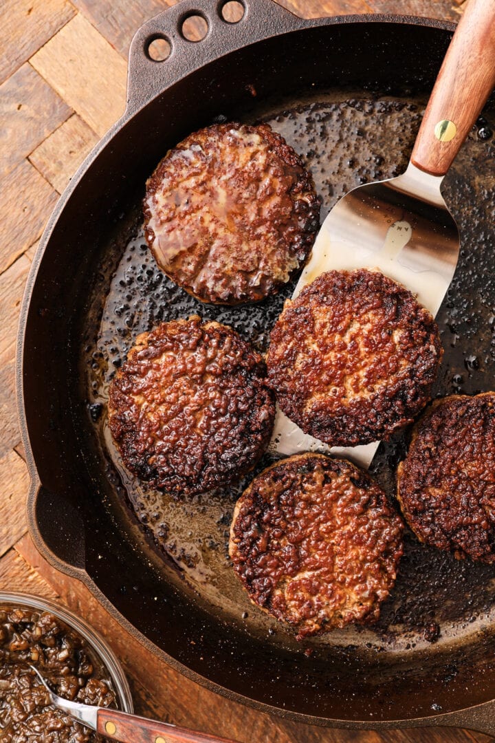 a cast iron skillet with 5 burger patties and a metal spatula. There is a small bowl of caramelized onions on the bottom left.