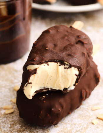 two chocolate covered ice cream bars that are stacked on top of each other. There is a plate of more ice cream bars off to the left and a small glass jar of chocolate dip.