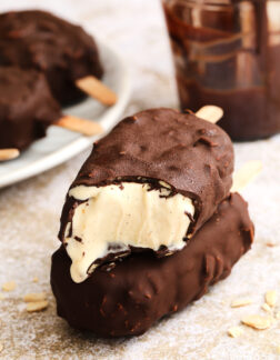 two chocolate covered ice cream bars that are stacked on top of each other. There is a plate of more ice cream bars off to the left and a small glass jar of chocolate dip.