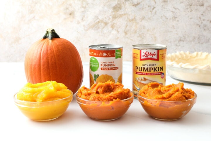 three small glass bowls of pumpkin puree, with cans of pumpkin and a small whole pumpkin behind the bowls in the background.