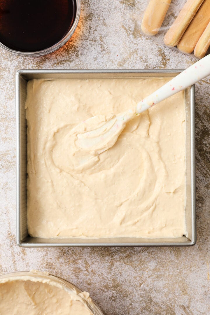 large dollop of coconut cream being spread over a layer of coffee soaked lady finger cookies, arranged in a metal square 9 x 9 pan.