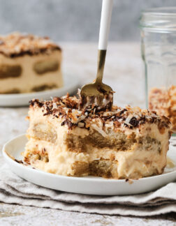 slice of coconut tiramisu, with a small fork sinking down into the slice to take a bite. There is a small jar of toasted coconut in the back right corner.