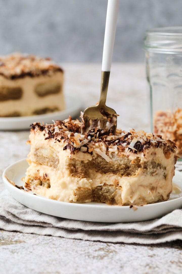 slice of coconut tiramisu, with a small fork sinking down into the slice to take a bite. There is a small jar of toasted coconut in the back right corner.