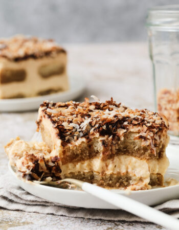 slice of coconut tiramisu, with a small fork sinking down into the slice to take a bite. There is a small jar of toasted coconut in the back right corner.