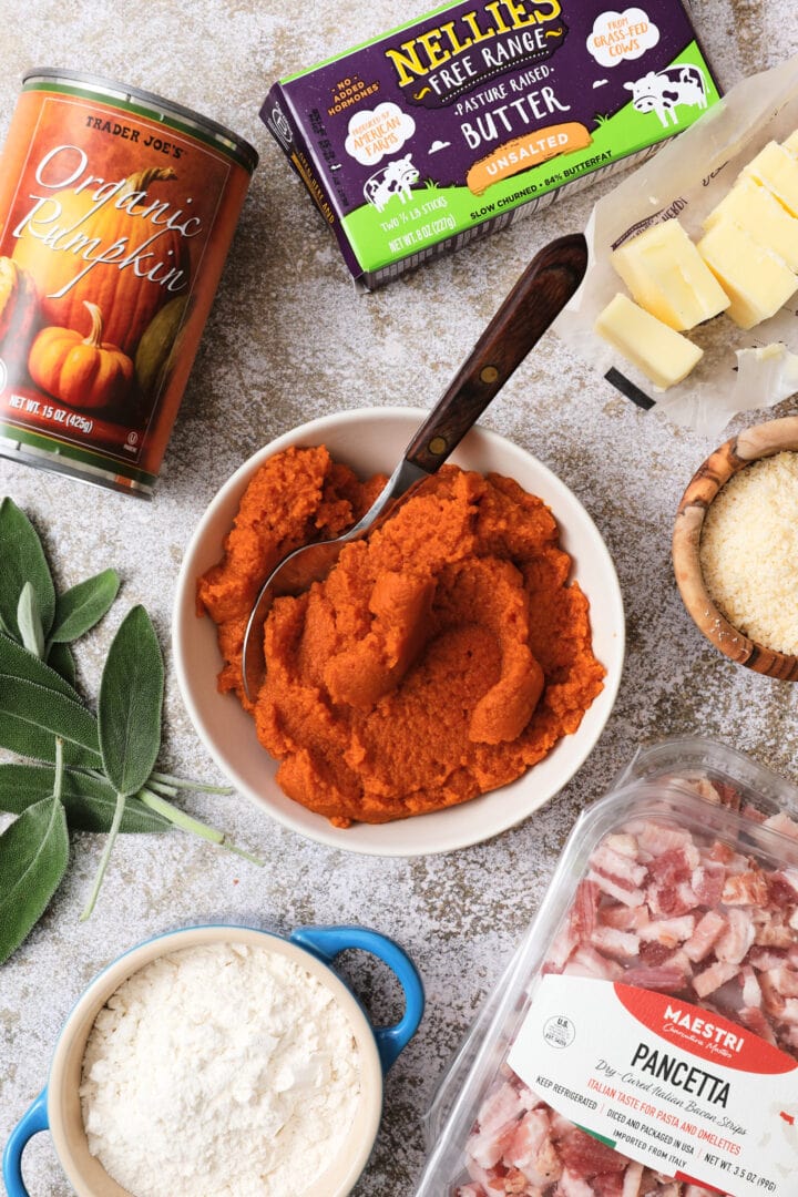 Assorted raw ingredients for making pumpkin gnocchi, including canned pumpkin, unsalted butter sticks, fresh sage leaves, pancetta, flour and parmesan cheese.