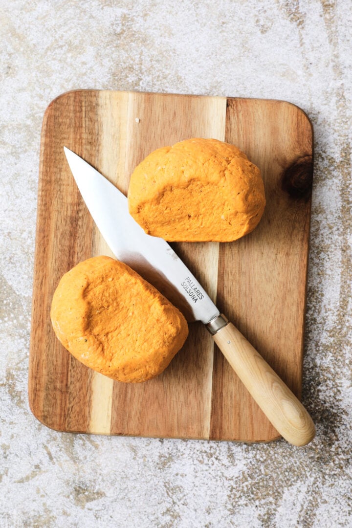 small wooden cutting board with two large pieces of pumpkin gnocchi dough and a large knife.