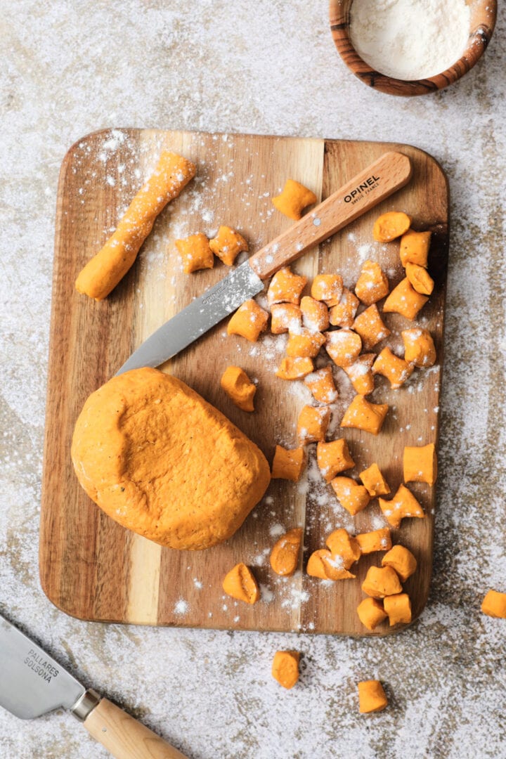 Small wooden cutting board that is dusted with flour and has shaped gnocchi pieces on it.