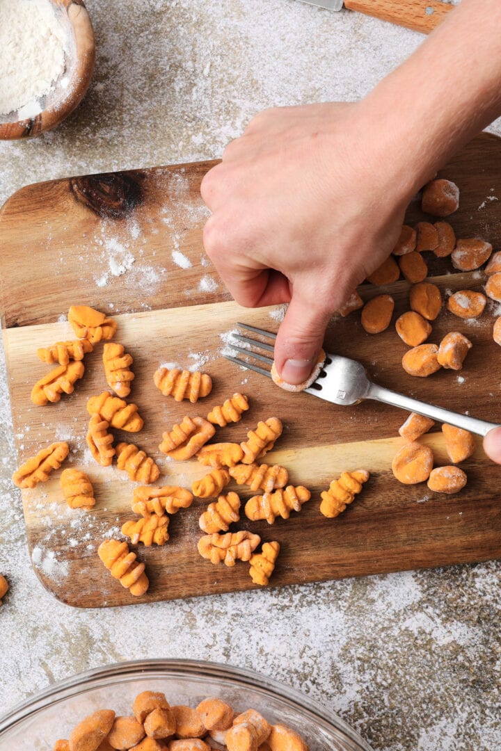 Small wooden cutting board that is dusted with flour and has shaped gnocchi pieces on it. There is someone's hands showing how to shape each noodle by using a fork.