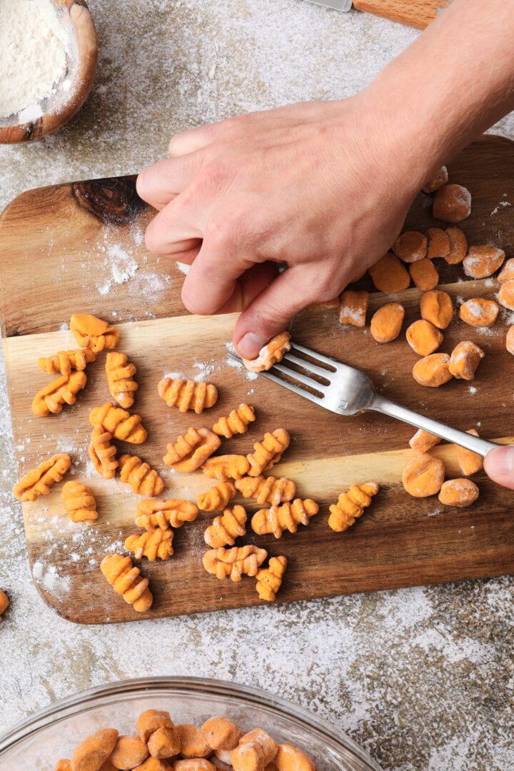 Small wooden cutting board that is dusted with flour and has shaped gnocchi pieces on it. There is someone's hands showing how to shape each noodle by using a fork.