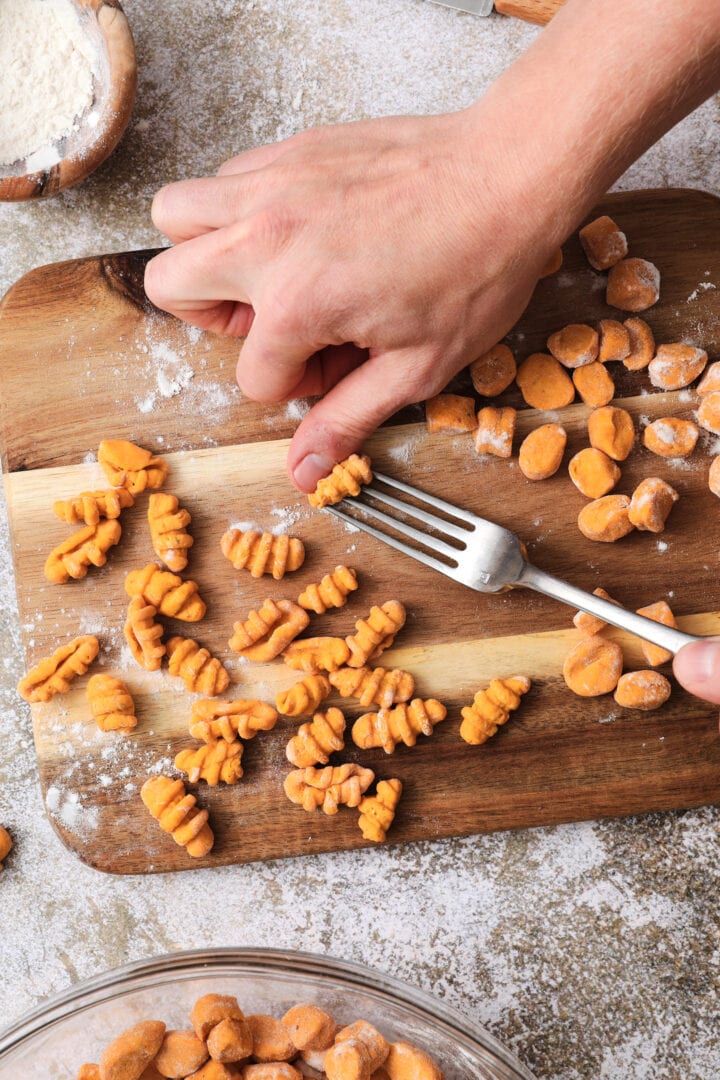 Small wooden cutting board that is dusted with flour and has shaped gnocchi pieces on it. There is someone's hands showing how to shape each noodle by using a fork.