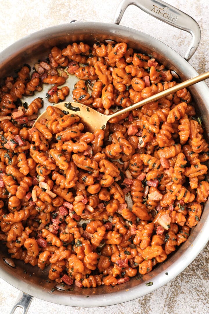 skillet of orange pumpkin gnocchi with a brown butter sauce, small pieces of pancetta and sage. There is a gold spoon on the side of the skillet.