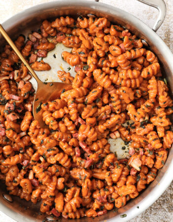 skillet of orange pumpkin gnocchi with a brown butter sauce, small pieces of pancetta and sage. There is a gold spoon on the side of the skillet.