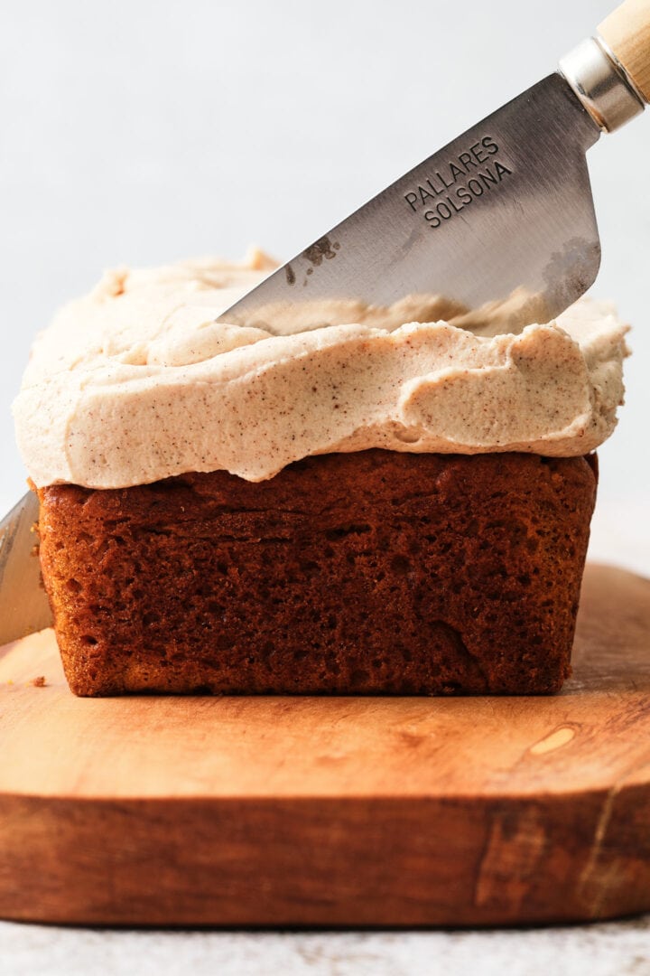 sliced pumpkin loaf resting on a wooden serving platter. There is a thick layer of frosting on the loaf.