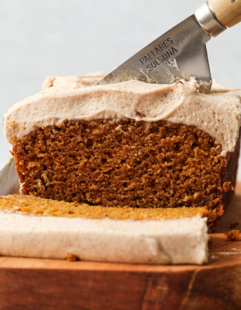 sliced pumpkin loaf resting on a wooden serving platter. There is a thick layer of frosting on the loaf.