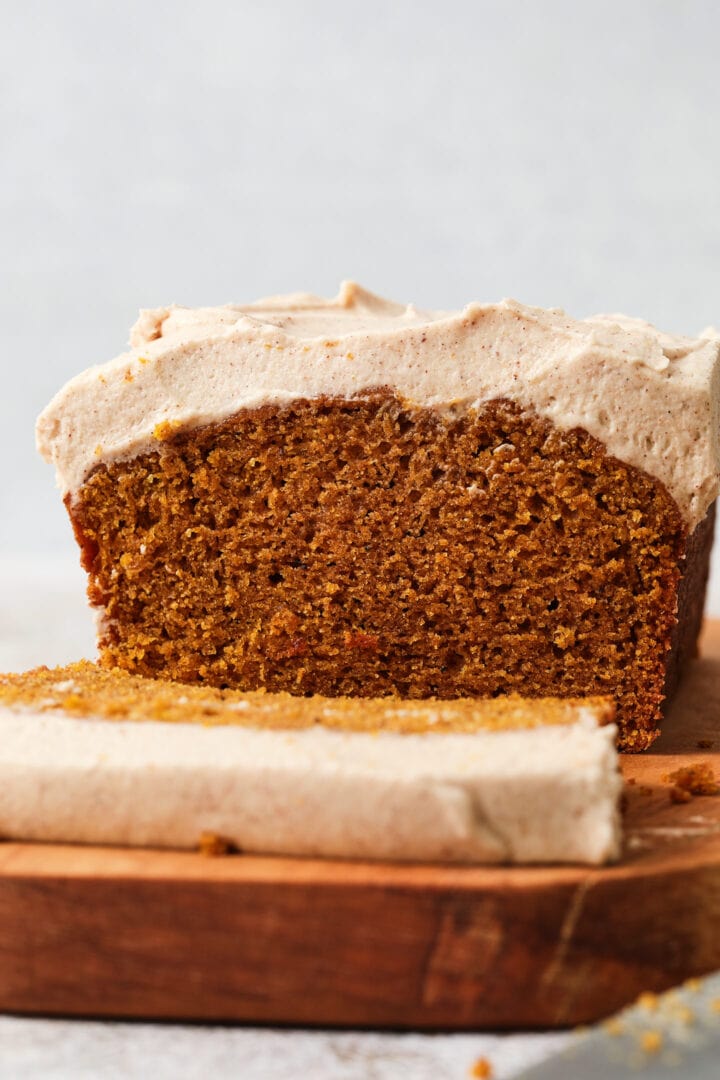 sliced pumpkin loaf resting on a wooden serving platter. There is a thick layer of frosting on the loaf.