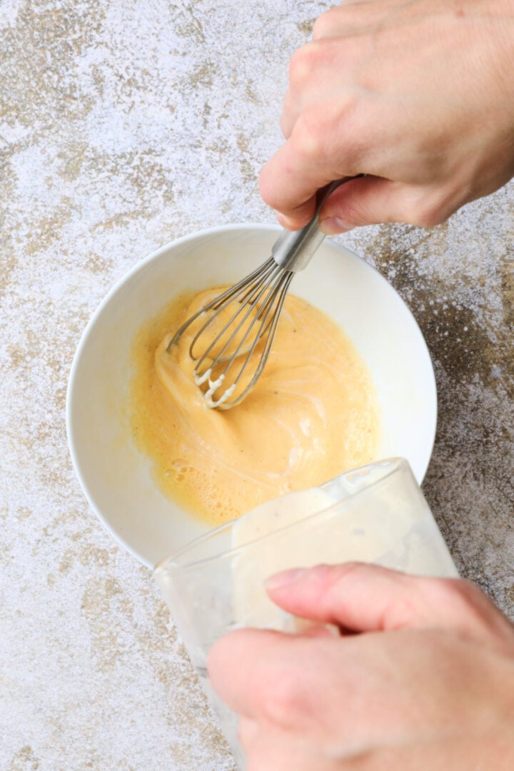 an egg being tempered by hot sauce in a small white bowl.