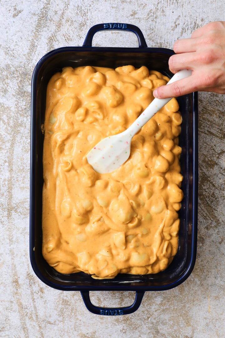 Unbaked blue pan of pumpkin macaroni and cheese, with a spatula that is smoothing the surface of the mac and cheese.