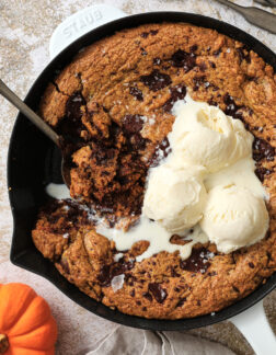 large pumpkin chocolate chip cookie with three scoops of vanilla ice cream on top. There is a large spoon taking a helping out of the cookie.