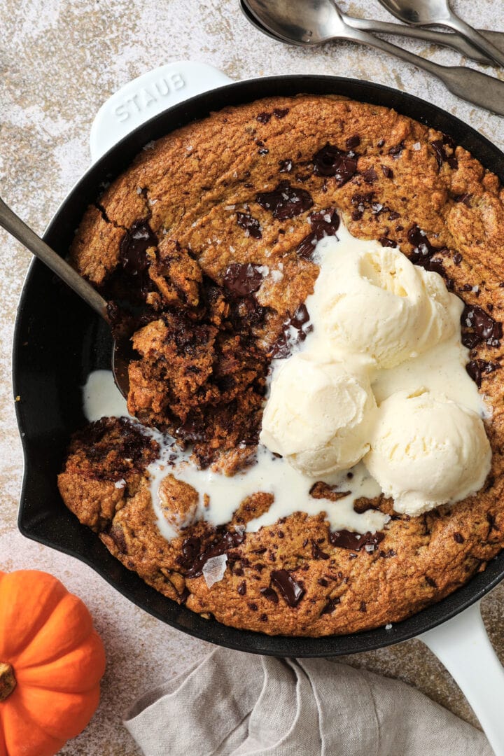 large pumpkin chocolate chip cookie with three scoops of vanilla ice cream on top. There is a large spoon taking a helping out of the cookie.