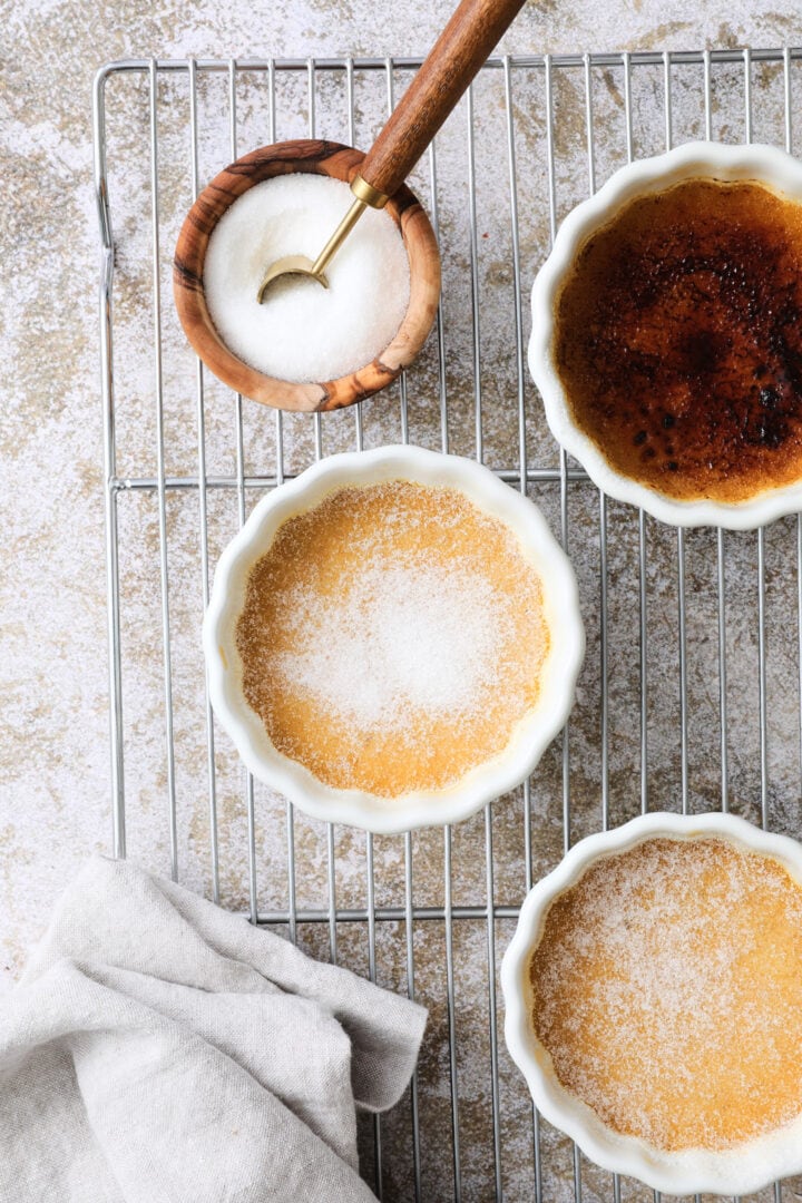 Three white ramekins of creme brulee arranged on a cooling rack. One of the ramekins has a burnt sugar crust, while the other two have not.