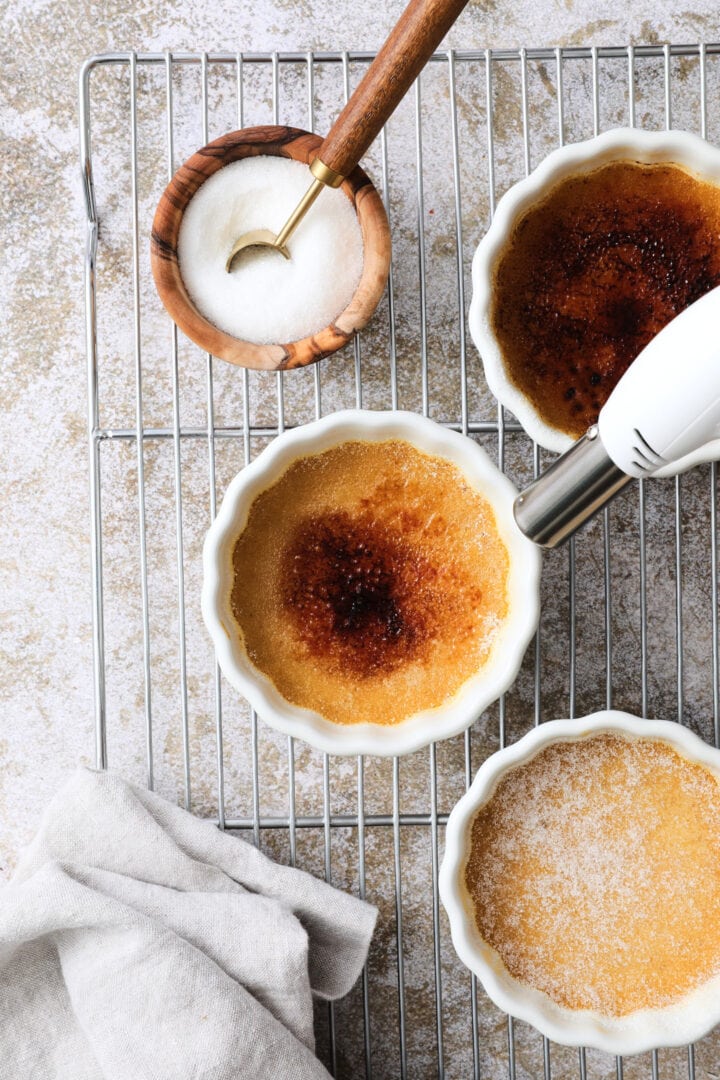 Three white ramekins of creme brulee arranged on a cooling rack. There is a white kitchen torch being used to burn the sugar on top of one of the ramekins.