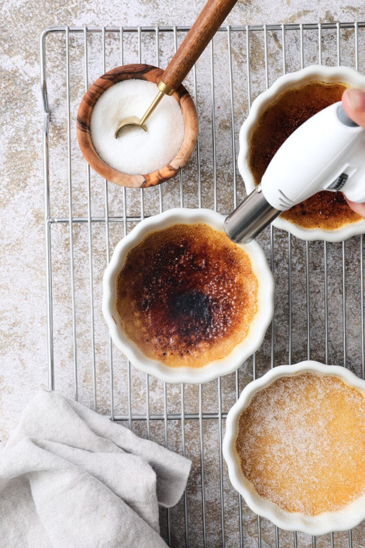 Three white ramekins of creme brulee arranged on a cooling rack. There is a white kitchen torch being used to burn the sugar on top of one of the ramekins.