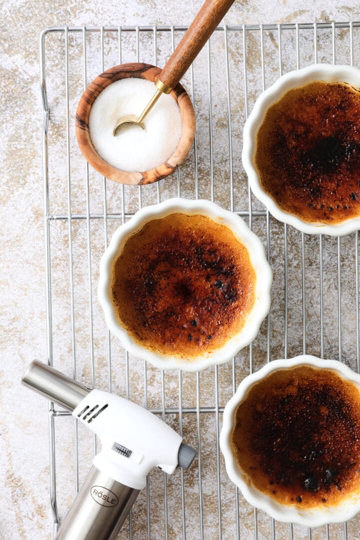 Three white ramekins of creme brulee arranged on a cooling rack. There is a white kitchen torch being used to burn the sugar on top of one of the ramekins.