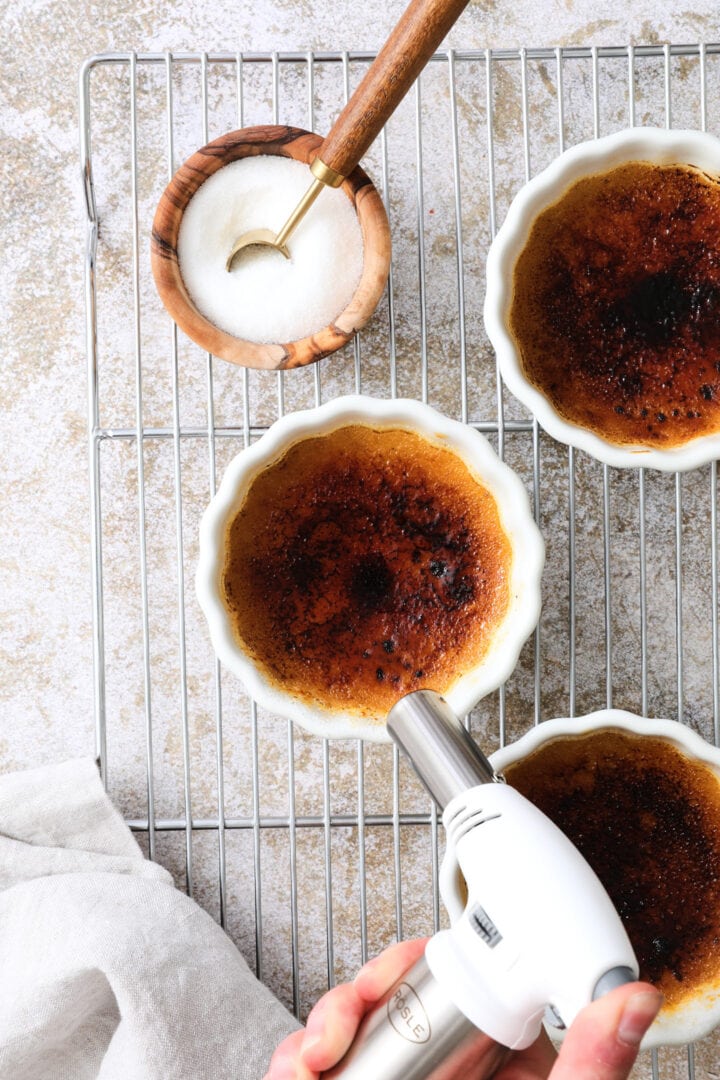 Three white ramekins of creme brulee arranged on a cooling rack. There is a white kitchen torch being used to burn the sugar on top of one of the ramekins.