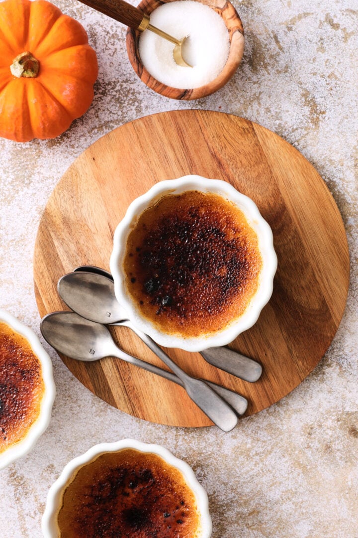 small white ramekin of pumpkin creme brulee, the burnt sugar crust has not been cracked yet, and there is a small orange pumpkin to the left hand side.