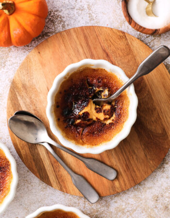 small white ramekin of pumpkin creme brulee, the burnt sugar crust has been cracked by a spoon and the pumpkin custard is just visible underneath.