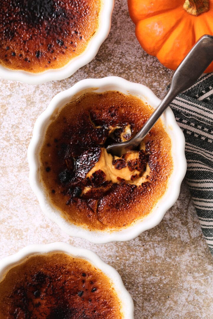 small white ramekin of pumpkin creme brulee, the burnt sugar crust has been cracked by a spoon and the pumpkin custard is just visible underneath.